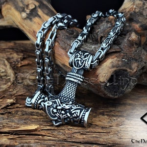 May include: A silver Mjolnir pendant necklace with intricate details and a braided chain. The pendant features a detailed design of the hammer head with a dragon head on the side. The chain is a box chain with a secure clasp.
