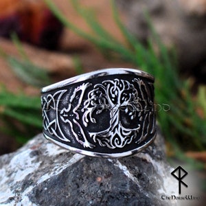 May include: A silver ring with a detailed design of a tree of life, surrounded by a Celtic knot and crescent moons. The ring is on a rocky surface.