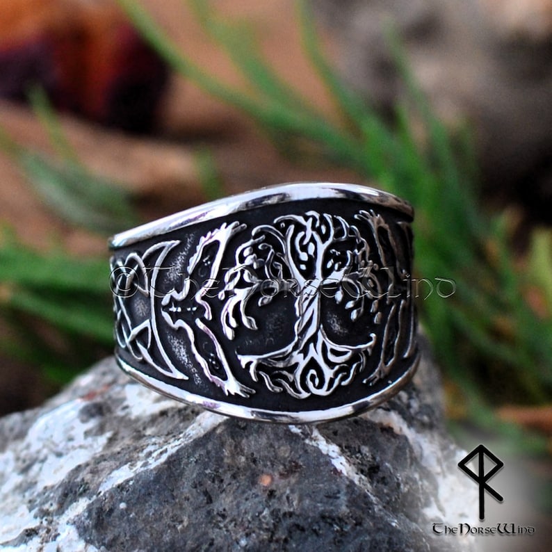 May include: A silver ring with a detailed design of a tree of life, surrounded by a Celtic knot and crescent moons. The ring is on a rocky surface.