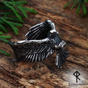 May include: A detailed silver ring featuring an eagle with outstretched wings. The ring is intricately designed with textured feathers and a detailed bird head. The ring is set against a rustic wooden background. The text "The Norse Wind" is visible.