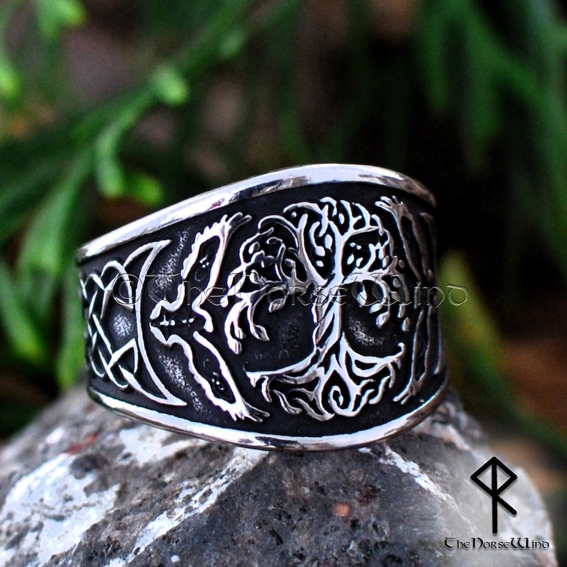 May include: A silver ring with a detailed design of a tree of life with a bird on a branch. The ring is wide and has a Celtic knot design on the side.
