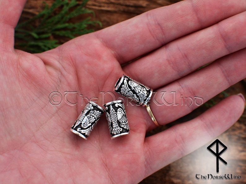 May include: Three silver hair beads with a detailed design of a bird with wings spread. The beads are on a hand, with a gold ring on the finger.