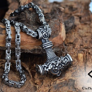May include: A silver Mjolnir pendant necklace with intricate knotwork designs and a braided chain. The pendant is on a wooden surface with a rune symbol in the background.