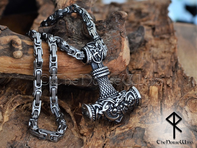 May include: A silver Mjolnir pendant necklace with intricate knotwork designs and a braided chain. The pendant is on a wooden surface with a rune symbol in the background.