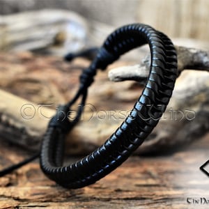 May include: Black leather bracelet with a braided design. The bracelet is tied with a knot and has a long, thin cord that can be adjusted to fit the wrist.