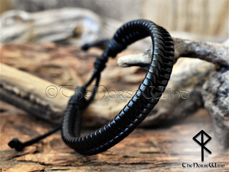 May include: Black leather bracelet with a braided design. The bracelet is tied with a knot and has a long, thin cord that can be adjusted to fit the wrist.