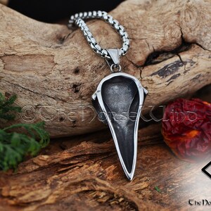 May include: A silver pendant in the shape of a raven's head, hanging from a silver chain. The pendant is detailed and has a sharp, pointed beak. The pendant is hanging against a wooden background.