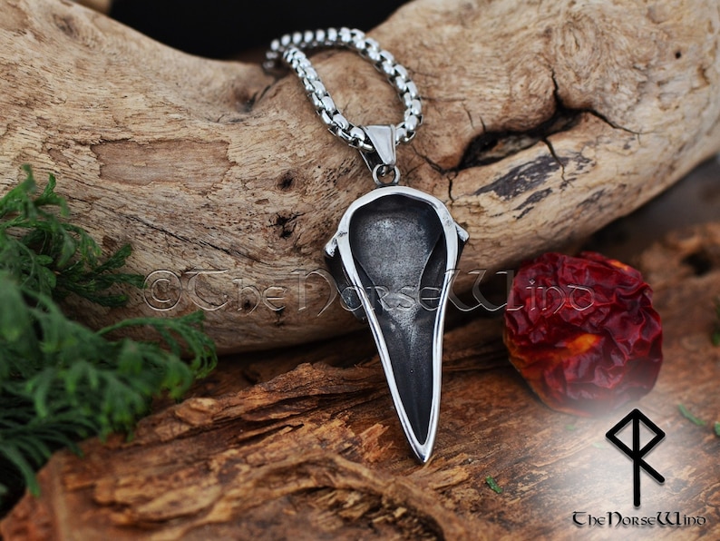 May include: A silver pendant in the shape of a raven's head, hanging from a silver chain. The pendant is detailed and has a sharp, pointed beak. The pendant is hanging against a wooden background.