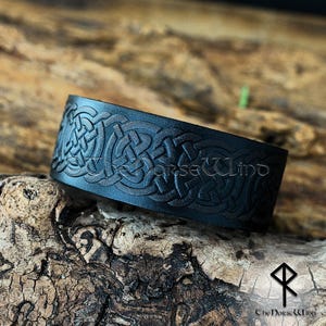 May include: A black leather cuff bracelet with an embossed Celtic knot design. The bracelet has the text "The Norse Wind" and a symbol. The cuff is shown on a textured wooden surface.
