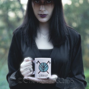 May include: A white ceramic mug with a woodgrain design features a blue and brown shield with crossed axes and the name "Lindsay".