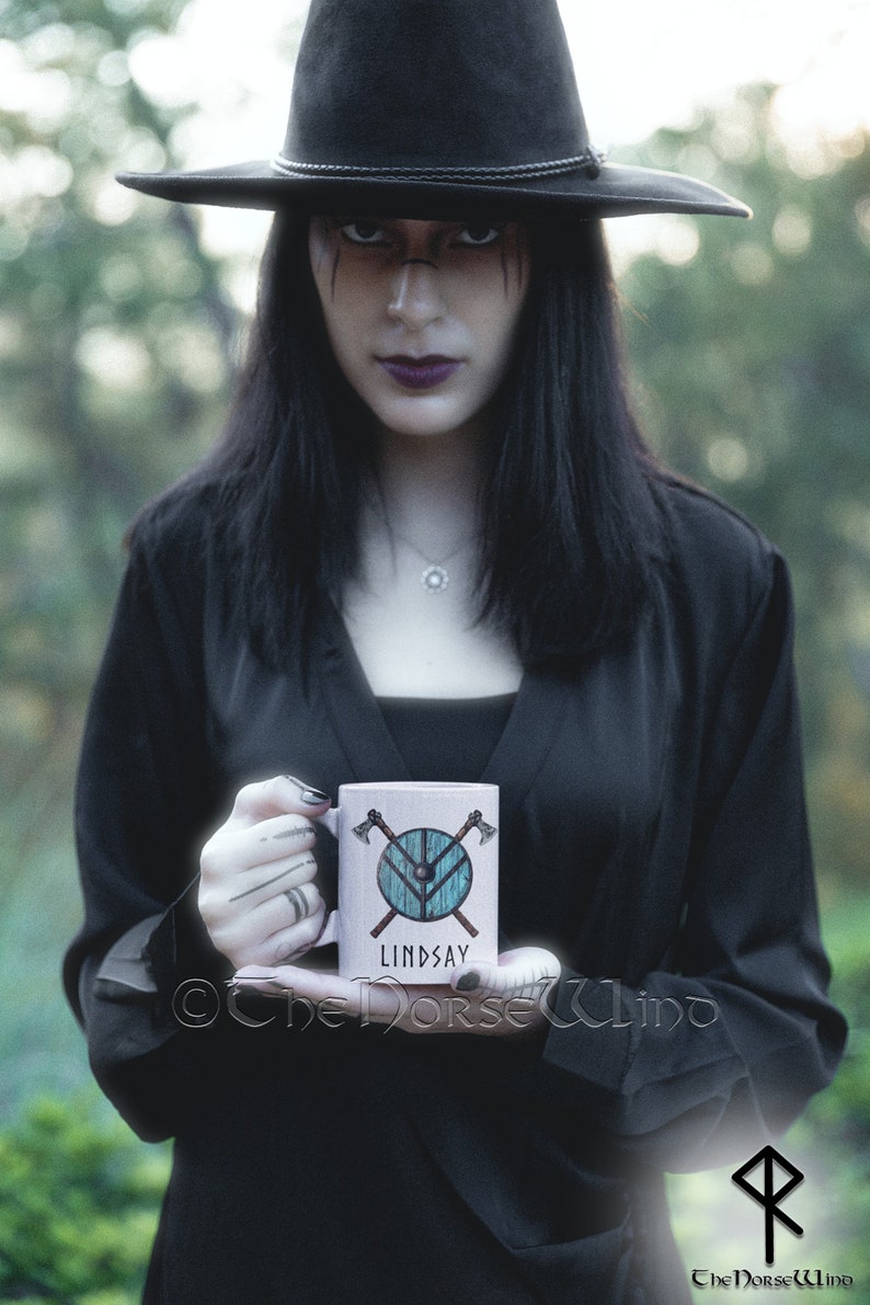 May include: A white ceramic mug with a woodgrain design features a blue and brown shield with crossed axes and the name "Lindsay".