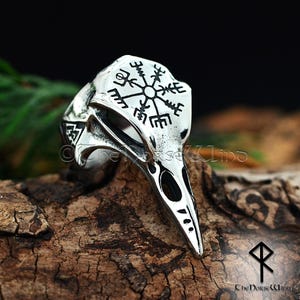 May include: A silver ring with a raven skull design and a Vegvisir compass symbol. The ring is made of stainless steel and has a black finish.