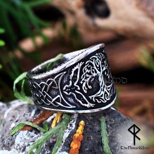 May include: A silver ring with a detailed design of a tree of life and a Celtic knot. The ring is wide and has a slightly curved shape.