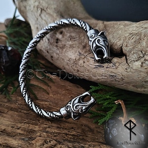 Op de afbeelding: Een zilverkleurige metalen armband met een gedraaid touwontwerp en twee gestileerde dierenkop-uiteinden. De armband is gegraveerd met de tekst The Norse Wind en bevat een logo met een geometrisch runensymbool aan de rechteronderkant.