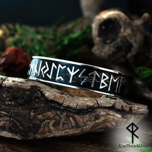 May include: A silver bracelet with a black enamel band and silver runes inscribed on the band. The bracelet is on a piece of wood.