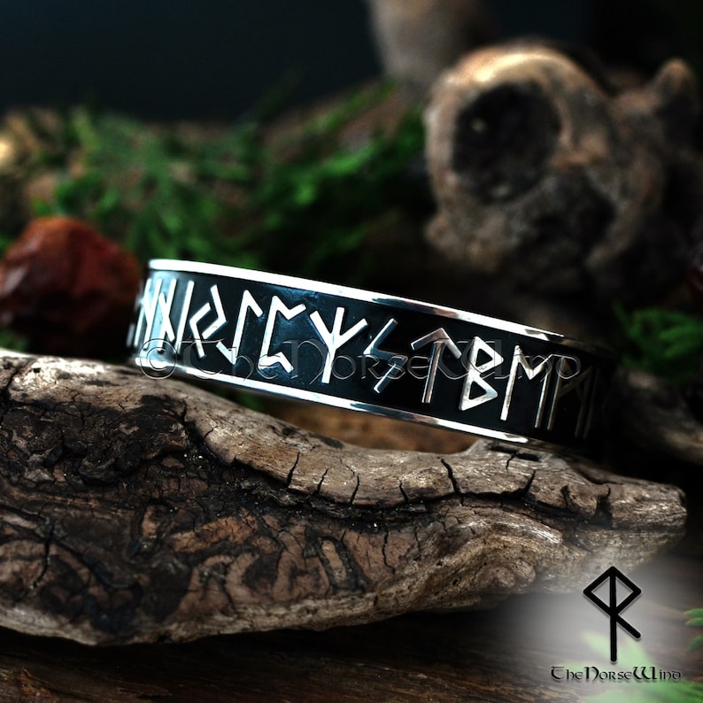 May include: A silver bracelet with a black enamel band and silver runes inscribed on the band. The bracelet is on a piece of wood.
