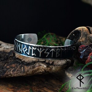 May include: A silver bracelet with a black background and engraved runes. The runes are in the style of the Elder Futhark, an ancient Germanic alphabet.