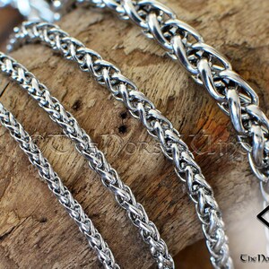 May include: Four silver braided chains of different widths, lying on a wooden surface. The chains are interlocked and appear to be made of metal.