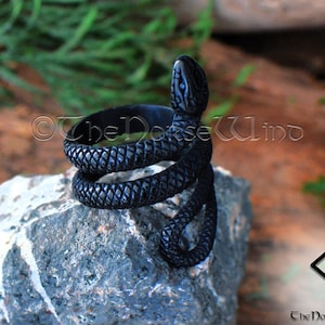 K&ouml;nnte beinhalten: Schwarzer Schlangenring mit strukturierter Oberfl&auml;che. Die Schlange ist um den Ringband gewickelt. Der Ring liegt auf einem grauen Stein.