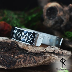 May include: A silver bracelet with a black band featuring the Vegvisir symbol, a Norse directional rune, in silver. The bracelet is on a wooden surface.