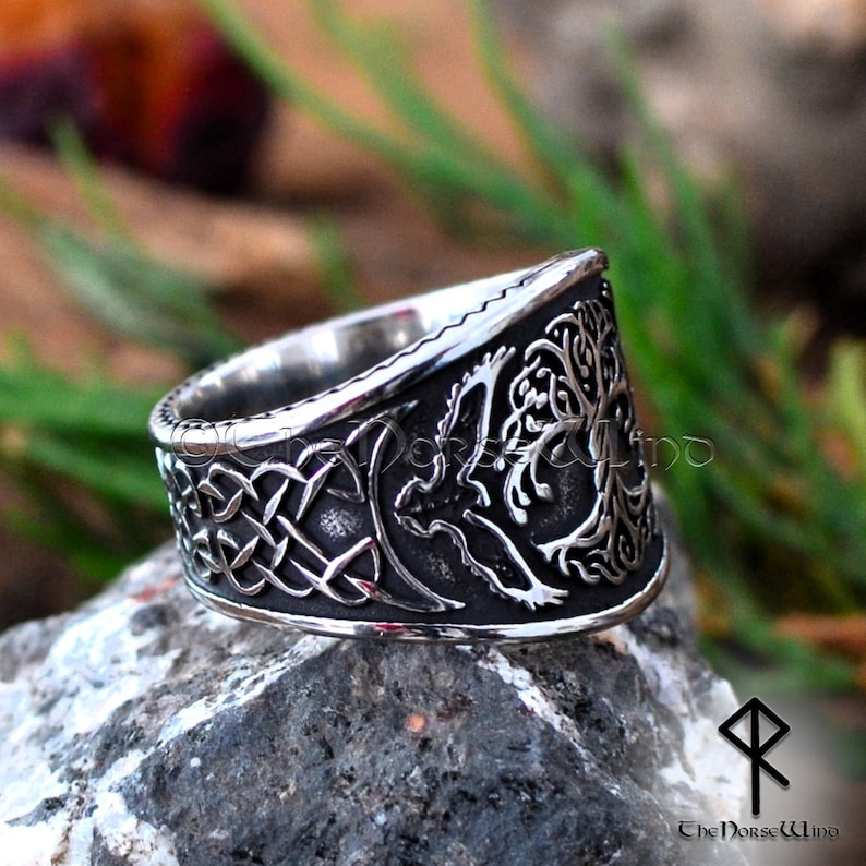 May include: A silver ring with a detailed design of a tree of life and a bird. The ring is wide and has a Celtic knot design on the band.