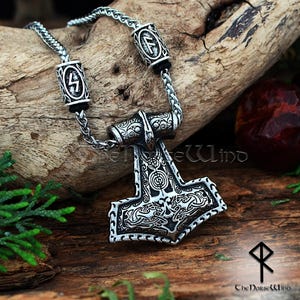 May include: A silver Mjolnir pendant necklace with intricate details and a chain with two silver beads featuring the Vegvisir symbol. The pendant is on a wooden surface.