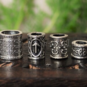 Könnte beinhalten: Vier silberne Haarperlen mit verschiedenen Designs. Die Perlen sind rund und haben ein erhabenes Design. Die Designs umfassen einen keltischen Knoten, eine Rune, einen Kreis mit Punkten und ein geflochtenes Muster.