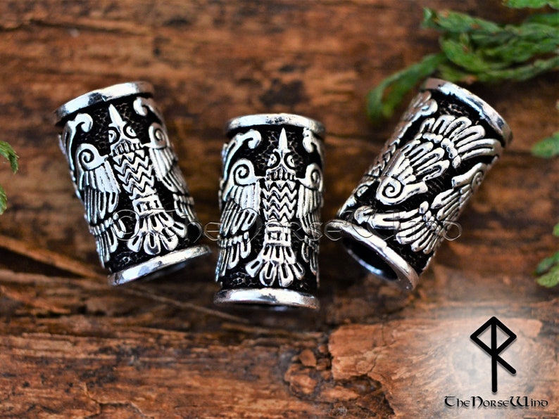 May include: Three silver-colored metal hair beads with a detailed design of a double-headed bird. The beads are on a wooden surface.