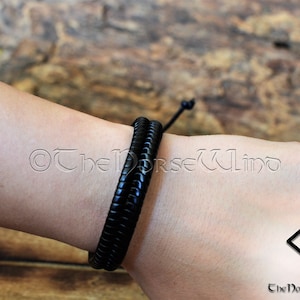 May include: A black leather bracelet with a braided design. The bracelet is tied with a knot and is worn on a person's wrist.