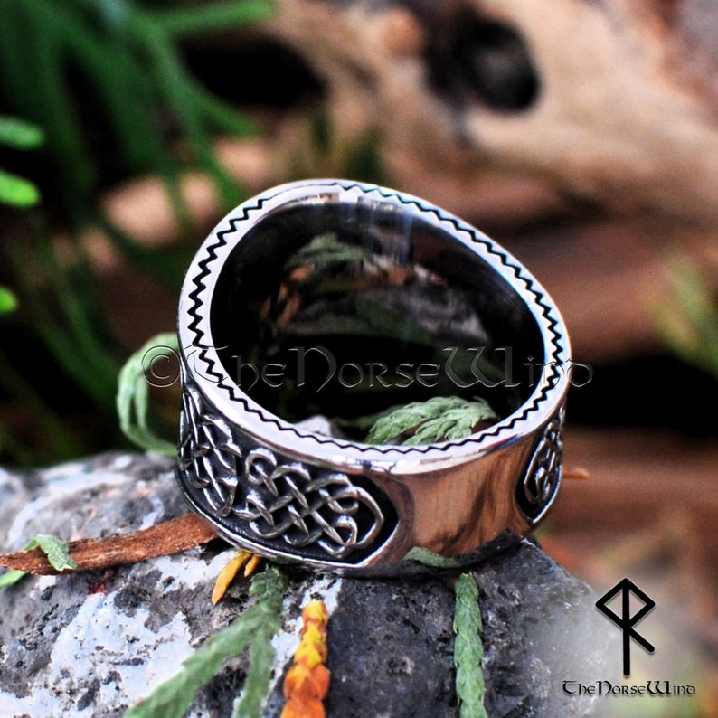 May include: A silver ring with a Celtic knot design. The ring is on a rock with green foliage in the background.