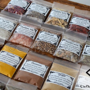 May include: A collection of small clear plastic bags filled with various herbs, spices, and salts. Each bag has a label with the name of the ingredient and its uses in magic and spell work. The labels are printed in black and white with a decorative border. The bags are arranged in rows on a wooden surface. The Norse Wind logo is visible in the bottom right corner.