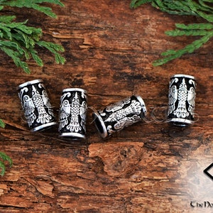May include: Four silver metal hair beads with a black background and a detailed design of a bird with its wings spread. The beads are on a wooden surface.