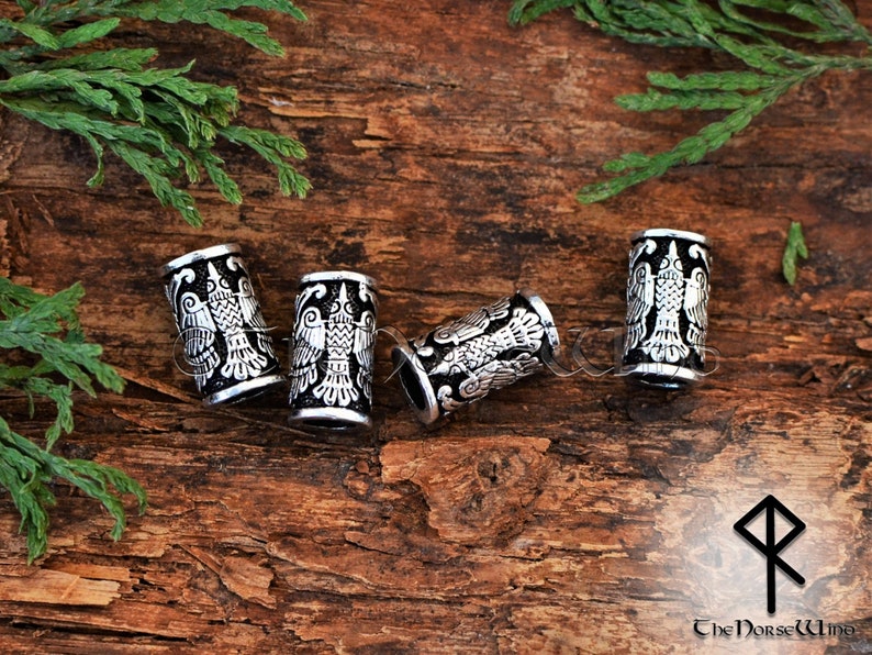 May include: Four silver metal hair beads with a black background and a detailed design of a bird with its wings spread. The beads are on a wooden surface.