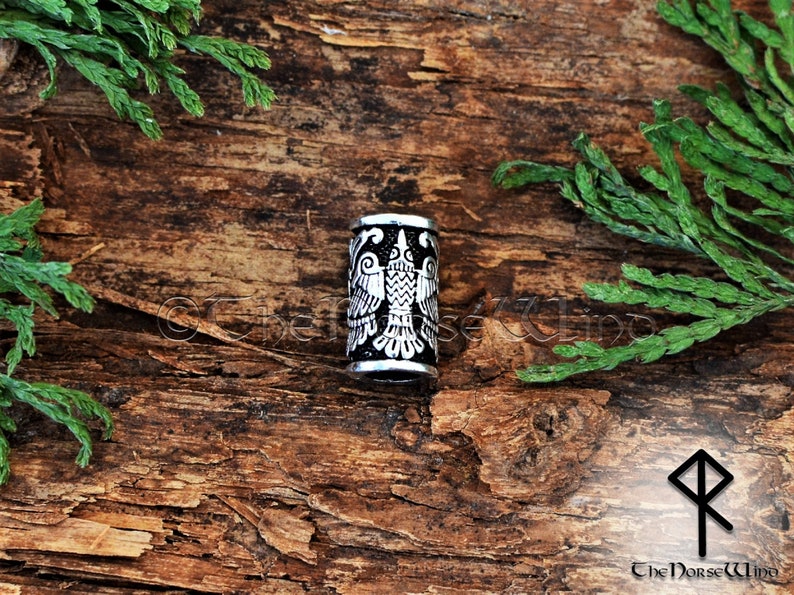 May include: A silver metal bead with a black background and a detailed design of a double-headed eagle. The bead is on a wooden surface.
