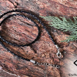 May include: A black braided leather cord necklace with silver clasps and a silver chain extension. The necklace is lying on a wooden surface.