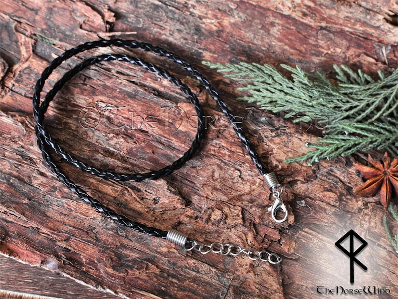 May include: A black braided leather cord necklace with silver clasps and a silver chain extension. The necklace is lying on a wooden surface.