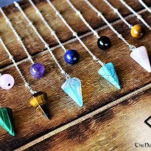 May include: A set of 10 crystal pendulums with silver chains. The pendulums are made of various crystals, including amethyst, rose quartz, tiger's eye, and turquoise. The pendulums are hanging from a wooden surface.
