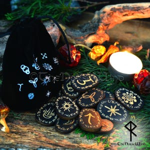Puede incluir: Un conjunto de 12 runas de madera negra con símbolos dorados, dispuestas sobre una superficie de madera. Las runas están en una bolsa de terciopelo negro con símbolos blancos. Las runas están rodeadas de follaje verde y una vela encendida.