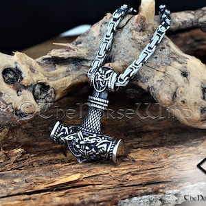 May include: A silver Mjolnir pendant necklace with intricate details, including a knotwork design and a small hammer head. The necklace is on a silver chain and is resting on a piece of driftwood.