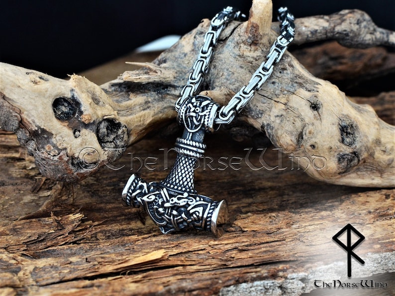 May include: A silver Mjolnir pendant necklace with intricate details, including a knotwork design and a small hammer head. The necklace is on a silver chain and is resting on a piece of driftwood.