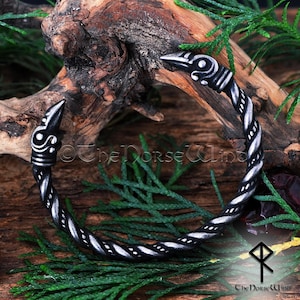 May include: A silver bracelet with a twisted rope design and a bird head on each end. The bracelet is on a wooden surface with green foliage.