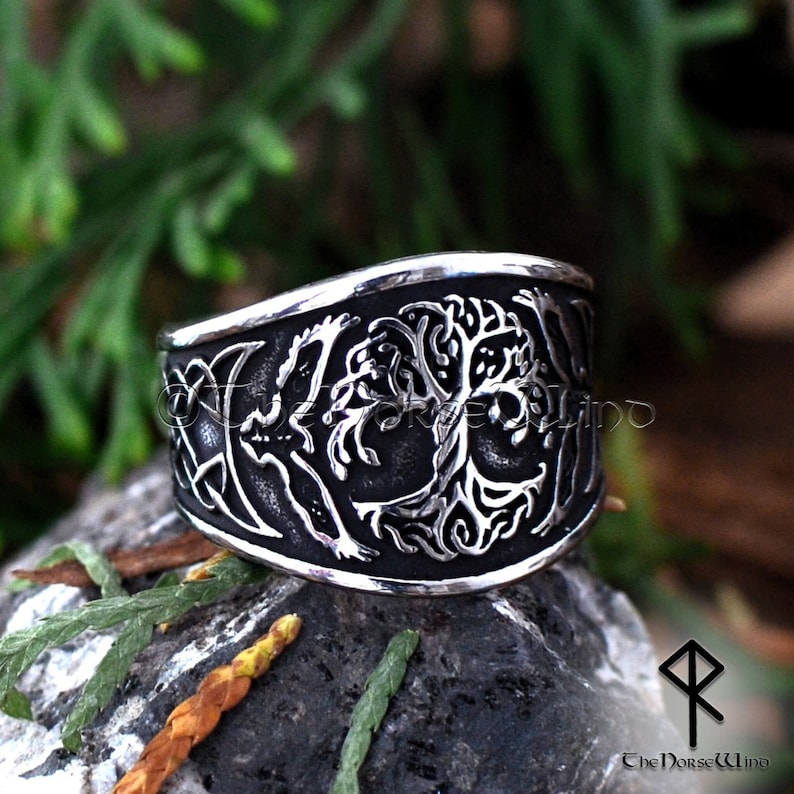 May include: A silver ring with a detailed design of a tree of life with a crescent moon and a bird on each side. The ring is on a dark gray rock.