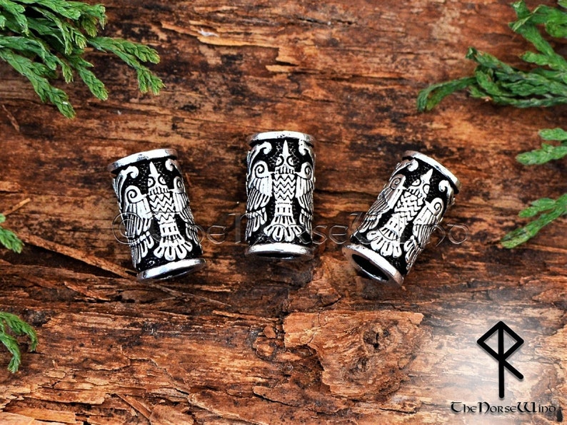 May include: Three silver-toned metal hair beads with a raised design of a double-headed eagle. The beads are on a wooden surface.