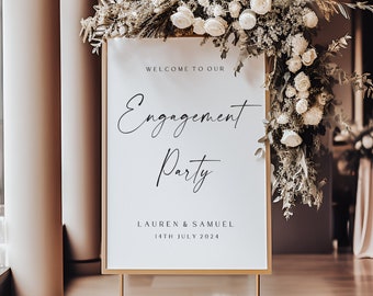May include: A white sign with gold trim on a gold stand with a floral arrangement behind it. The sign reads "Welcome to our Engagement Party Lauren & Samuel 14th July 2024".