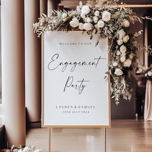 May include: A white sign with gold trim on a gold stand with a floral arrangement behind it. The sign reads "Welcome to our Engagement Party Lauren & Samuel 14th July 2024".