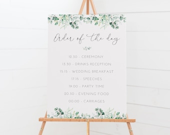 Puede incluir: Un cartel blanco con un borde de follaje verde en un caballete. El cartel dice "Orden del día" y enumera el horario del día de la boda: 12:30 Ceremonia, 13:30 Recepción de bebidas, 15:15 Desayuno de boda, 17:15 Discursos, 19:00 Hora de la fiesta, 20:30 Cena, 00:00 Carruajes.