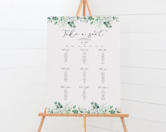 May include: A wedding seating chart with a green leafy border. The chart is titled "Take a Seat" and lists guest names by table number. The chart is displayed on a wooden easel.