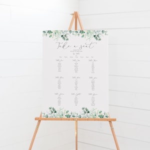 May include: A wedding seating chart with a green leafy border. The chart is titled "Take a Seat" and lists guest names by table number. The chart is displayed on a wooden easel.