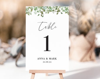 May include: White table number card with green eucalyptus leaves and the text "Table 1 Anna & Mark 24.08.24" on a wooden easel.