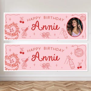 May include: A light pink rectangular birthday banner featuring the text Happy Birthday Annie in red script. The design includes red line art of a heart-shaped cake with the number 21, cherries, a cocktail glass, a disco ball, and a champagne bottle.